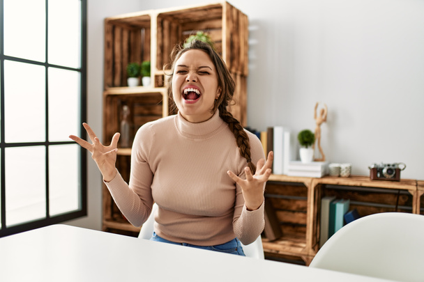 Fiatal spanyol lány alkalmi ruhát visel ül az asztalon otthon őrült és őrült kiabálás és kiabálás agresszív arckifejezéssel és felemelt karokkal. frusztráció fogalma.  - Fotó, kép