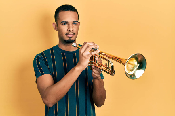Young african american man playing trumpet relaxed with serious expression on face. simple and natural looking at the camera.  - 写真・画像
