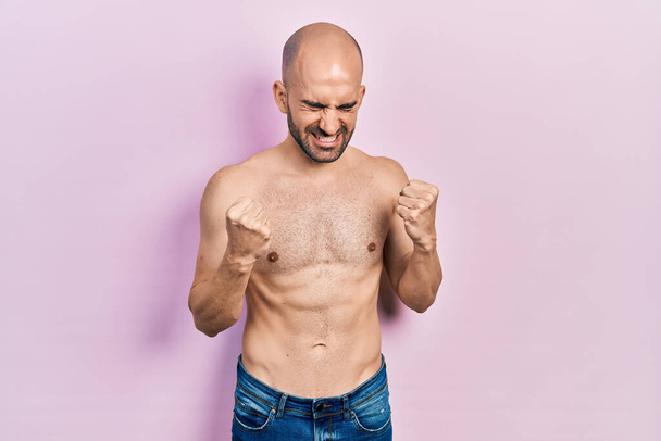 Young bald man standing shirtless very happy and excited doing winner gesture with arms raised, smiling and screaming for success. celebration concept.  - Fotoğraf, Görsel