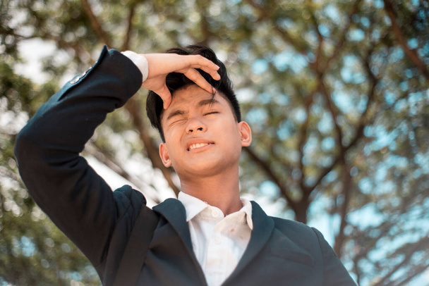 Um jovem estudante asiático ausente cerra sua testa em frustração, possivelmente esquecendo algo ou falhou um assunto. No parque ou campus. - Foto, Imagem