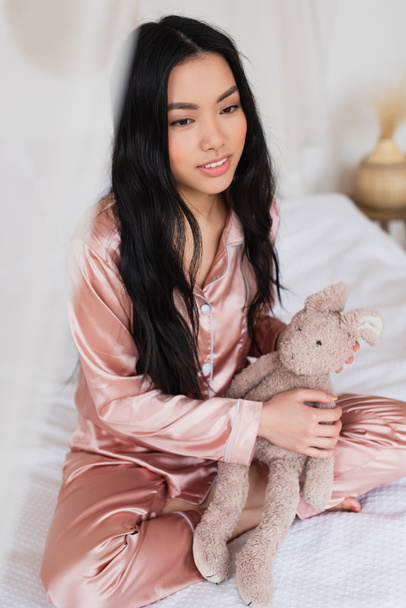 junge Asiatin im rosa Seidenpyjama sitzt mit überkreuzten Beinen auf dem Bett und hält Stofftier im Schlafzimmer - Foto, Bild