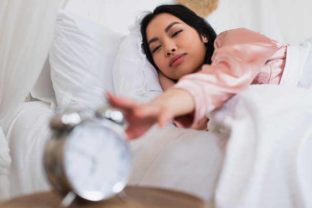 Uykulu genç Asyalı kadın yatakta uzanıyor ve yatak odasındaki komodinin üzerindeki çalar saate uzanıyor. - Fotoğraf, Görsel