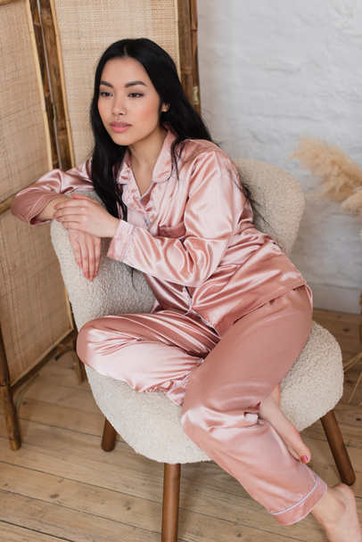 dromerige jonge aziatische vrouw in roze zijde pyjama zitten op stoel in slaapkamer - Foto, afbeelding