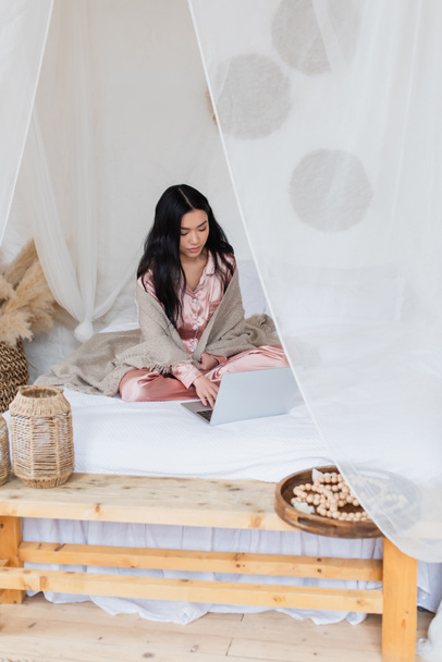 ernsthafte junge asiatische Frau in seidenem Pyjama und Decke sitzt mit überkreuzten Beinen auf dem Bett und tippt auf dem Laptop im Schlafzimmer - Foto, Bild