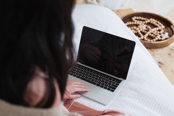 καλλιεργημένη άποψη της νεαρής γυναίκας που κάθεται στο κρεβάτι και πληκτρολογεί σε φορητό υπολογιστή στο υπνοδωμάτιο - Φωτογραφία, εικόνα