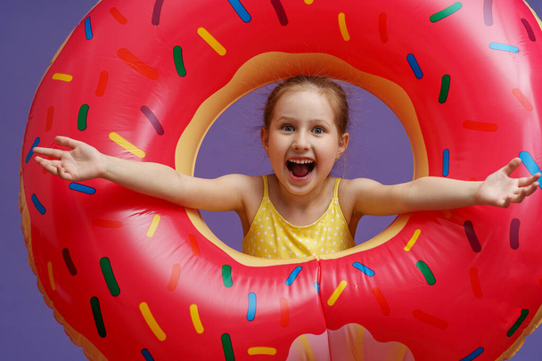 Happy emotional little girl in a swimsuit with an inflatable circle in the shape of a donut. children's beach accessories. summer children's weekend. sun baths. summer vacation trip to the sea. - Foto, Imagen