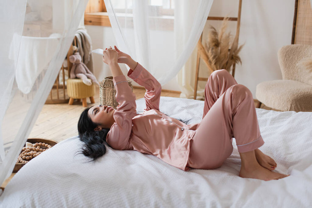 fiatal ázsiai nő selyem pizsamában fekszik az ágyon, és kezében mobiltelefon a hálószobában - Fotó, kép