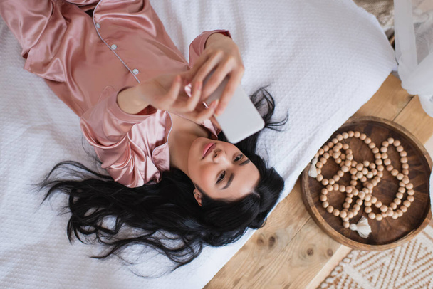 top view of young asian woman in silk pajamas lying on bed and taking selfie with cellphone in bedroom - Photo, Image