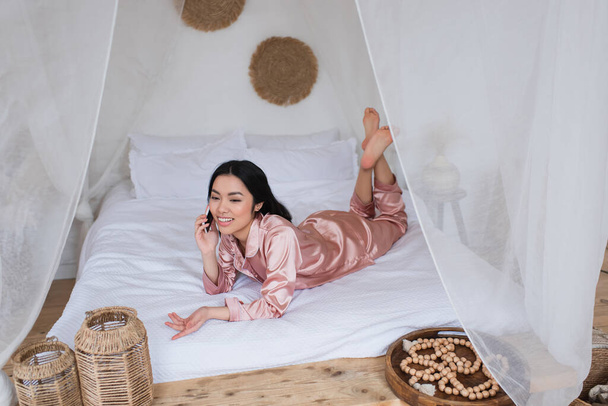 joven asiático mujer en rosa seda pijama acostado en cama y hablando en celular en dormitorio - Foto, Imagen