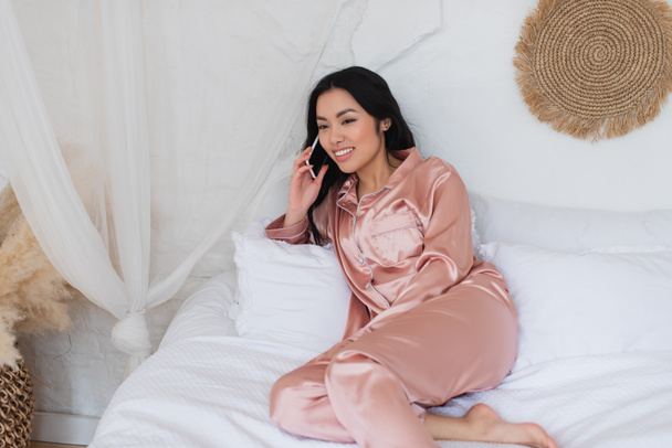 šťastný mladý asijské žena v růžové hedvábí pyžamo ležící na posteli a mluvení na mobilu v ložnici - Fotografie, Obrázek