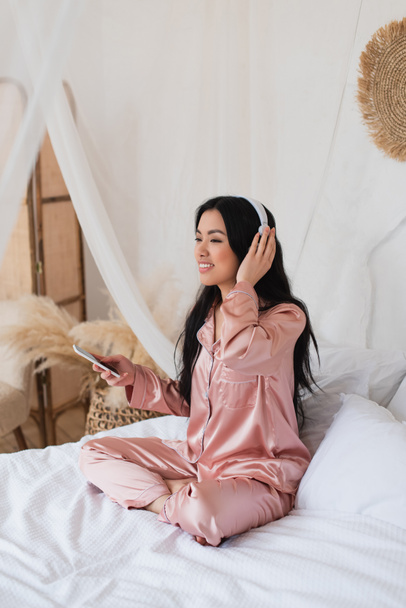 young asian woman in pink silk pajamas sitting on bed with cellphone and touching headphones with hand in bedroom - Photo, Image
