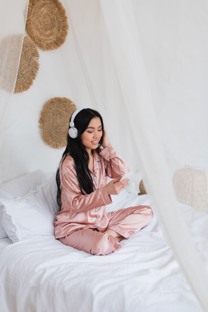 pozitív fiatal ázsiai nő ül az ágyon fejhallgatóval és nézi mobiltelefon a hálószobában - Fotó, kép