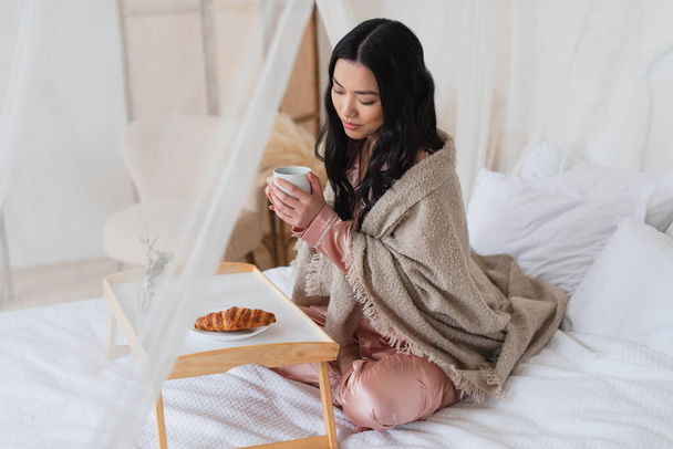 νεαρή Ασιάτισσα με μεταξωτές πιτζάμες και κουβέρτα που κάθεται στο κρεβάτι με σταυρωμένα πόδια και τρώει πρωινό στην κρεβατοκάμαρα - Φωτογραφία, εικόνα