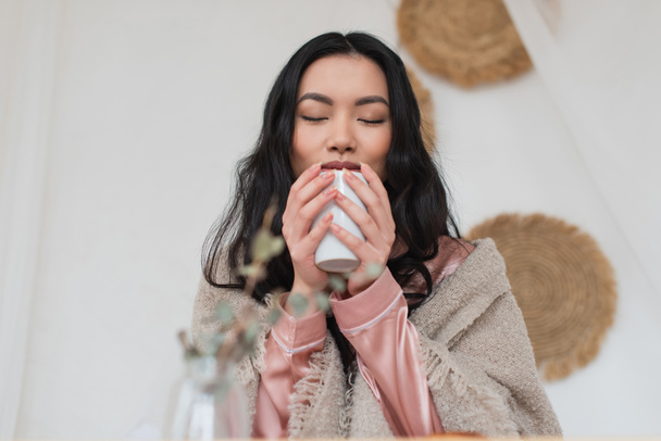 νεαρή Ασιάτισσα που κρατάει ένα φλιτζάνι καφέ στα χέρια και απολαμβάνει το άρωμα στην κρεβατοκάμαρα - Φωτογραφία, εικόνα