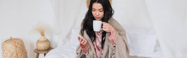 νεαρή Ασιάτισσα με μεταξωτές πιτζάμες και κουβέρτα που κάθεται στο κρεβάτι, πίνοντας καφέ και κρατώντας κινητό στην κρεβατοκάμαρα, πανό - Φωτογραφία, εικόνα