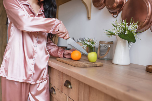 μερική άποψη της νεαρής γυναίκας σε ροζ μεταξωτές πιτζάμες κοπής φρούτων στην κουζίνα - Φωτογραφία, εικόνα