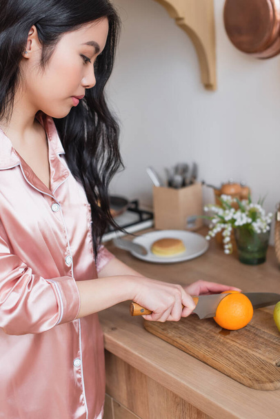 young asian woman in pink silk pajamas cutting orange in kitchen - Photo, Image