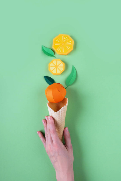 Hnd holding waffle cone with paper fruits on green background. Orange icecream concept - Fotó, kép