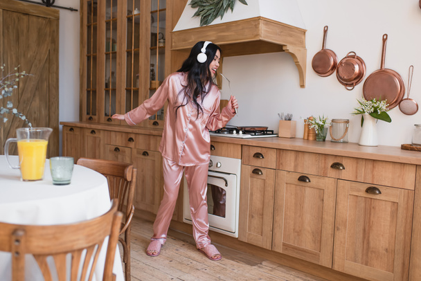 młoda azjatycka kobieta w różowej jedwabnej piżamie i słuchawki gotowanie śniadanie i śpiew z łopatką w kuchni - Zdjęcie, obraz
