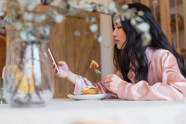 sorrindo jovem asiático mulher no pijama de seda comer panquecas com garfo e olhando para o celular na cozinha - Foto, Imagem