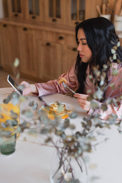 positive young asian woman in silk pajamas eating pancakes with fork and looking at cellphone with blank screen in kitchen - Fotografie, Obrázek