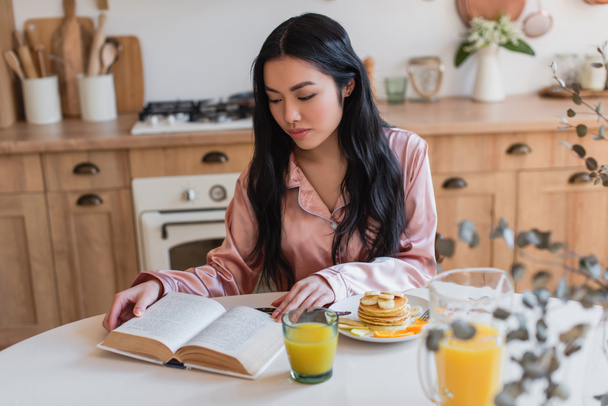Nuori aasialainen tyttö vaaleanpunainen silkki pyjama istuu pöydällä ja lukeminen kirja kun ottaa aamiaisen keittiössä - Valokuva, kuva