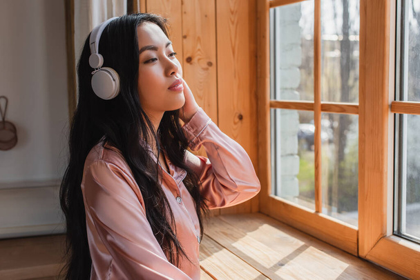 rêveuse jeune femme asiatique en pyjama de soie debout près de la fenêtre avec seuil avec écouteurs et tenant la main près de la tête à la maison - Photo, image