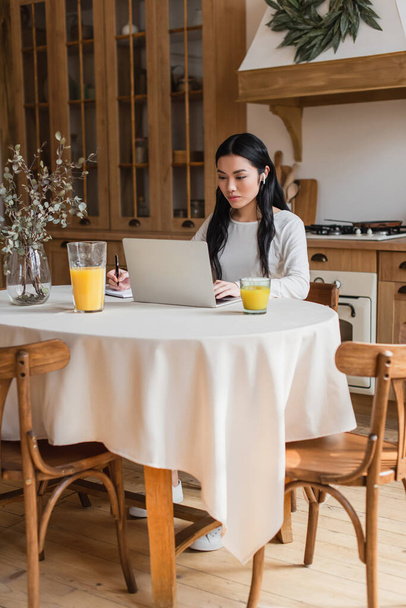 serious young asian woman in earphones sitting on table, using laptop and writing with pen in notebook in kitchen - Photo, Image