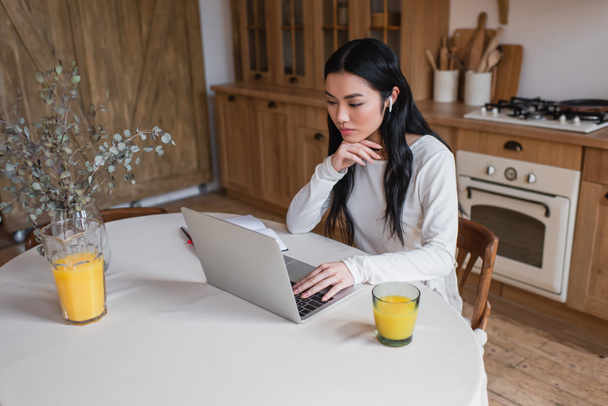 młoda azjatycka kobieta w słuchawki siedzi na stole z laptopem i trzyma rękę blisko twarzy w kuchni - Zdjęcie, obraz