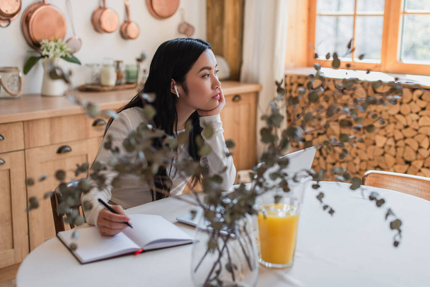 Kulaklıklı rüya gibi genç Asyalı kadın mutfaktaki kalem, defter ve dizüstü bilgisayarla masada oturuyor. - Fotoğraf, Görsel