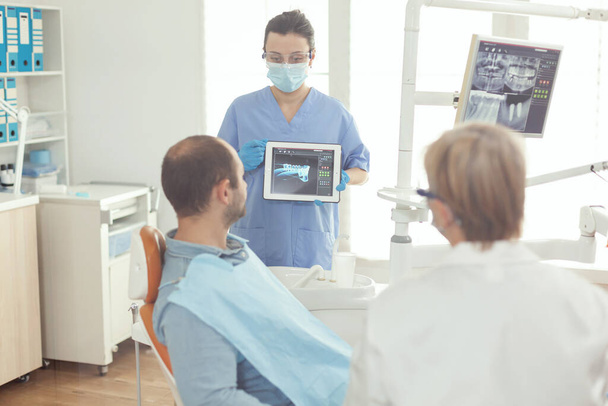 Медсестра-стоматолог указывает на цифровой экран, объясняя рентген зуба больному человеку - Фото, изображение