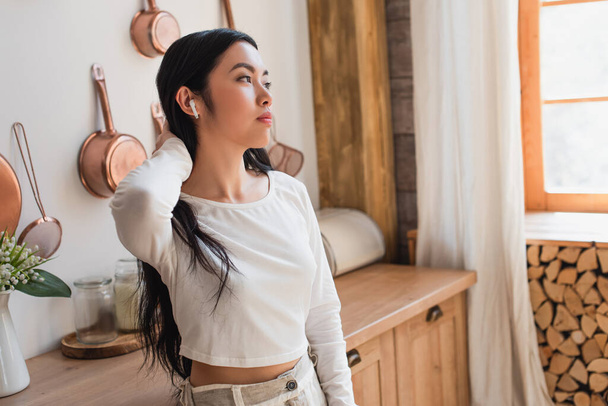 cansado joven asiático mujer en blanco blusa de pie con auriculares y celebración de la mano cerca del cuello en la cocina - Foto, imagen