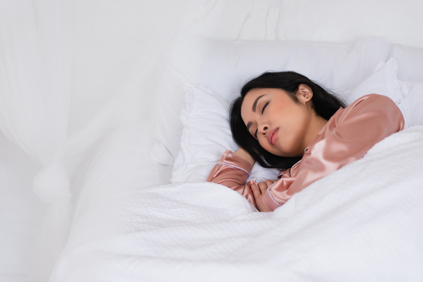 junge asiatische Frau im seidenen Pyjama schlafend im Bett mit weißer Bettwäsche im Schlafzimmer - Foto, Bild