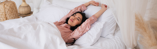 young asian woman in silk pajamas waking up with outstretched hands and closed eyes in bedroom, banner - Foto, Bild