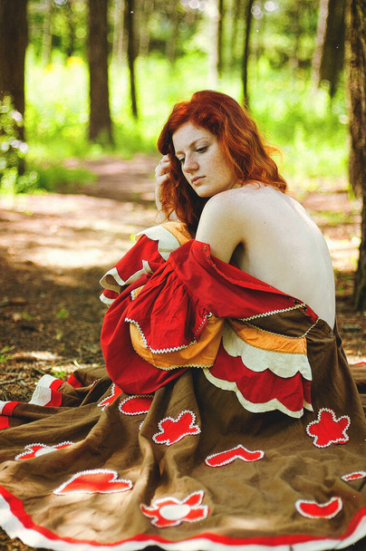 Świetna dziewczyna z piegami i rudymi włosami, pozująca z opuszczoną czerwoną sukienką i gołymi ramionami na dzikiej przyrodzie. Portret boho kobiety pozującej na letniej wsi. Szczęśliwy styl życia mody.  - Zdjęcie, obraz