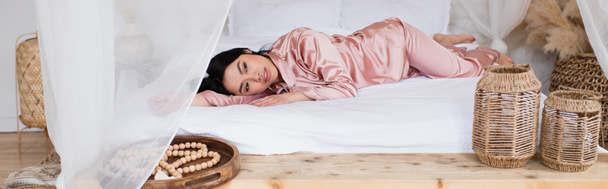 mujer asiática joven en pijama de seda acostada con la mano extendida sobre ropa blanca en el dormitorio, pancarta - Foto, Imagen