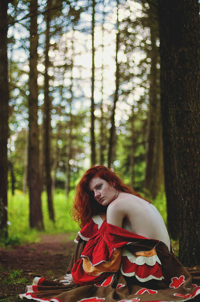 Девушка с рыжими волосами позирует на дикой природе в нарядном красном платье и голых плечах. Портрет женщины-бохо, позирующей в летней сельской местности. Счастливый образ жизни.  - Фото, изображение
