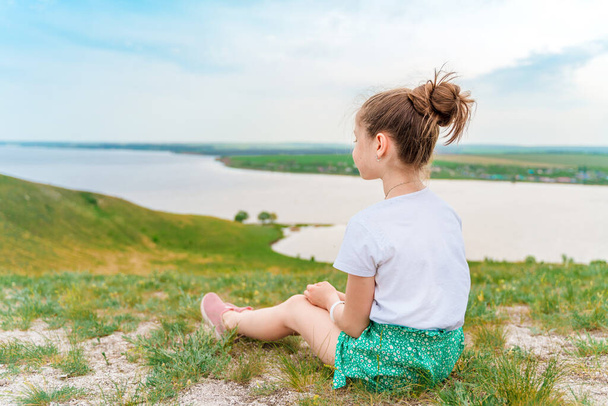 Rear view of a little girl child sitting on a green meadow with a delightful view of the hills and the river - Fotoğraf, Görsel