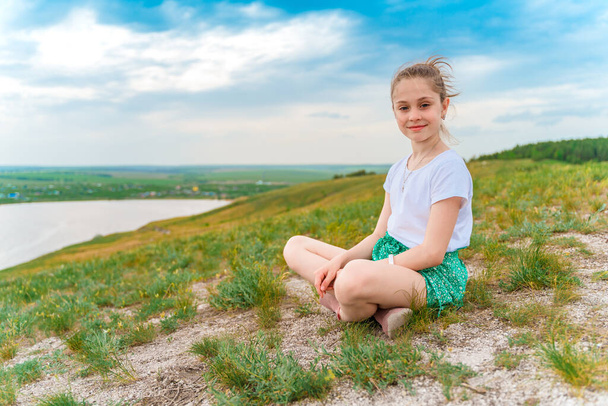 Visão traseira de uma criança menina sentada em um prado verde com uma vista encantadora das colinas e do rio - Foto, Imagem