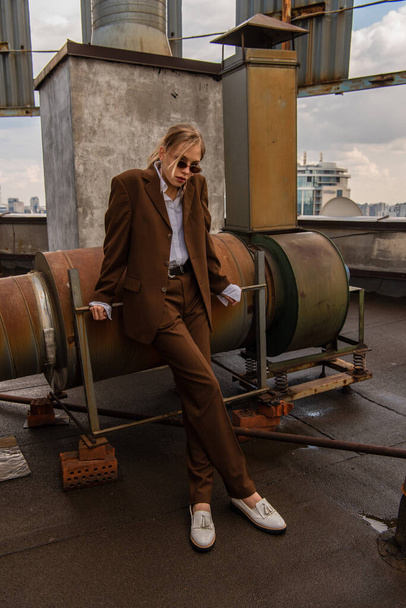 full length of blonde model in sunglasses and trendy brown suit posing on rooftop  - Фото, изображение