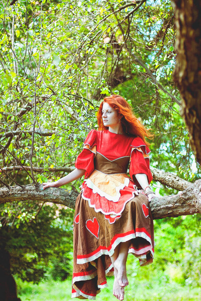 Chica con estilo con pecas y el pelo rojo en vestido largo rojo sosteniendo flor de amapola roja en el campo verde en la naturaleza. Retrato de mujer boho posando en el campo de verano. Feliz estilo de vida. Momento atmosférico.  - Foto, Imagen