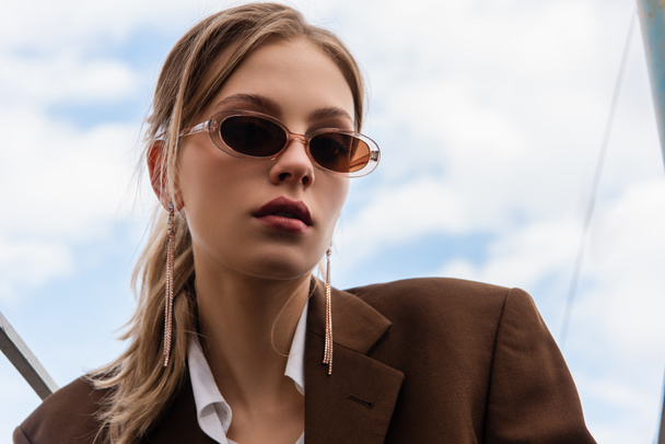young woman in sunglasses and blazer posing against blue sky - 写真・画像
