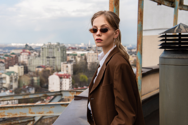 young model in stylish sunglasses posing on rooftop with cityscape on blurred background - Photo, Image