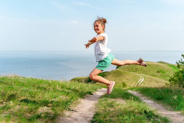 Een klein meisje kind springt vrolijk in de lucht op een pad in het midden van groene weiden - Foto, afbeelding