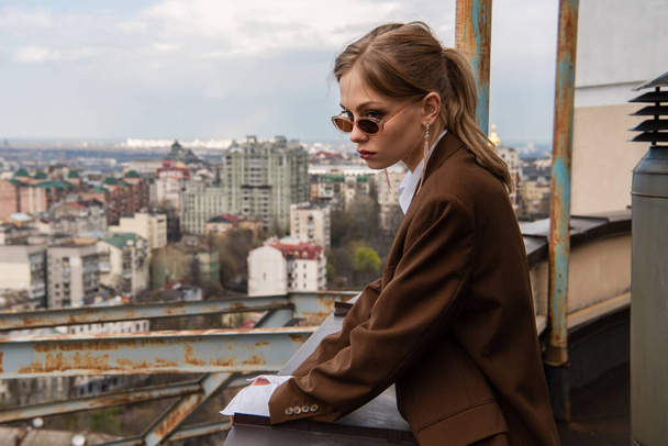 thoughtful model in stylish sunglasses posing on rooftop with cityscape on blurred background - Valokuva, kuva