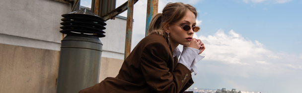 thoughtful model in stylish sunglasses posing on rooftop with cityscape on blurred background, banner - Photo, Image