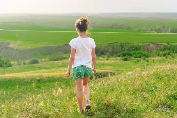  Οπίσθια άποψη κοριτσάκι περπάτημα μέσα από λιβάδια και περπάτημα κατηφόρα, καλοκαίρι όμορφο τοπίο - Φωτογραφία, εικόνα
