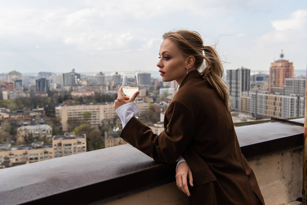 joven mujer sosteniendo copa de vino blanco y posando con paisaje urbano sobre fondo borroso - Foto, imagen