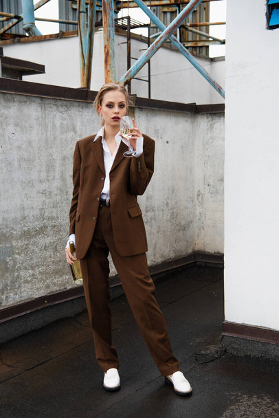 full length of young woman in stylish suit holding bottle and drinking wine on rooftop - Foto, Imagem