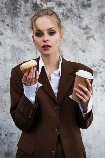 young woman in stylish outfit holding paper cup and doughnut against concrete wall - Φωτογραφία, εικόνα